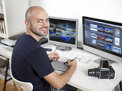 Ein Mann sitzt an einem Schreibtisch mit zwei Computermonitoren, einem Grafiktablett und einer Kamera.